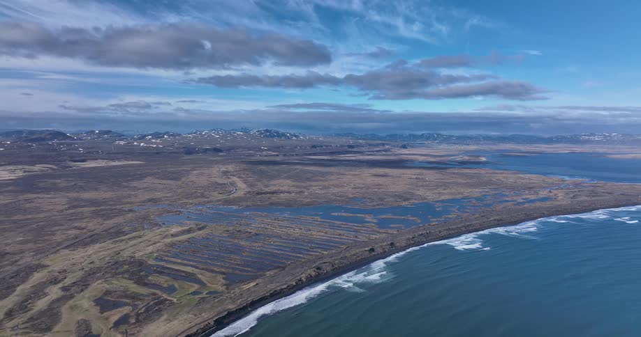 Static high-angle aerial shot of a vast coastal landscape featuring wetlands, a dark sand beach, and distant snow-capped mountains