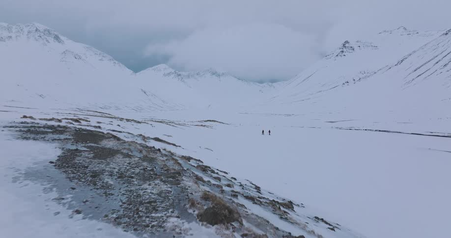 A slow-moving aerial shot reveals two distant figures hiking across a vast, desolate, snow-covered valley in Iceland