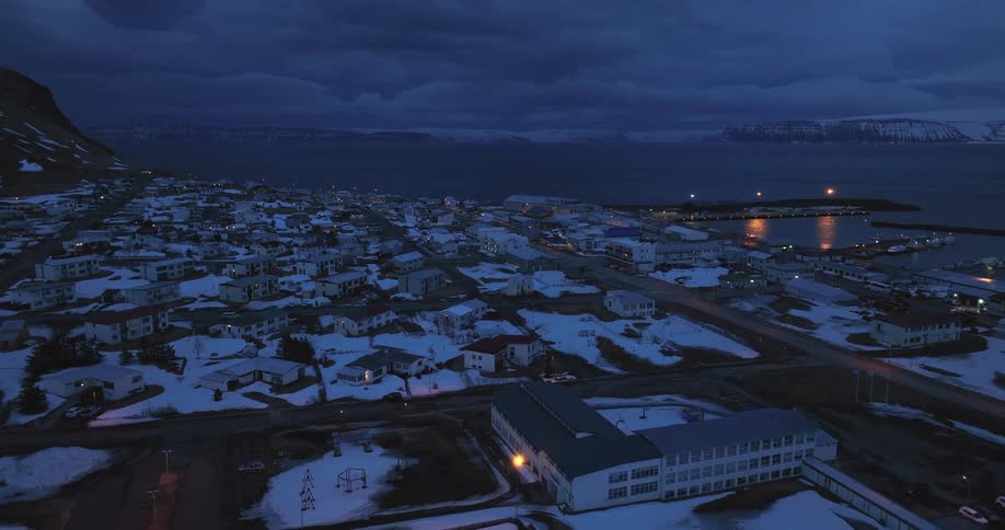 A slow pulling back aerial reveals a snow-dusted seaside village at twilight, with glowing lights dotting the landscape