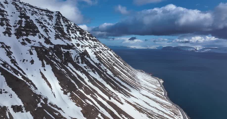 A static aerial view captures the dramatic landscape of a snow-covered mountain plunging into the ocean along a rugged coastline