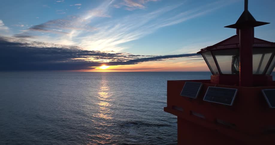 An aerial view slowly approaches a red lighthouse as the sun rises over the tranquil ocean