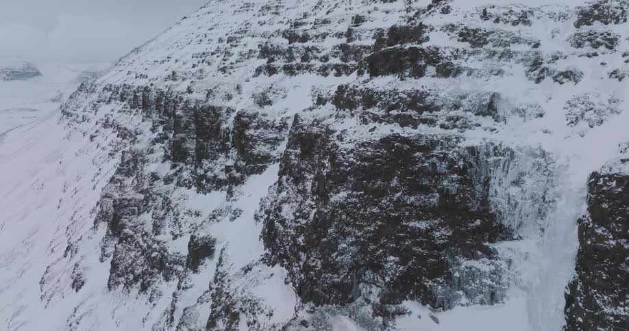 A static aerial shot captures snow and ice cascading down a rugged, frozen mountain face next to a massive icefall