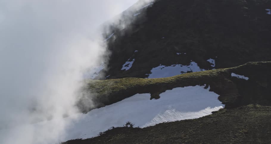Thick fog slowly rolls over a mossy mountainside with patches of snow, gradually revealing the rugged landscape
