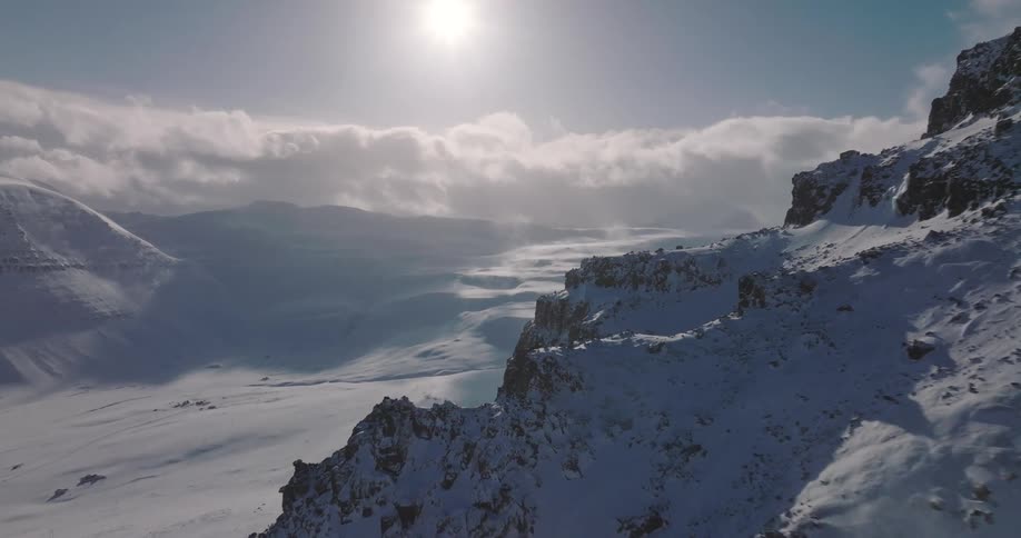 An epic aerial shot flies alongside a rugged, snow-covered mountain ridge as strong winds blow snow across the vast, frozen landscape below