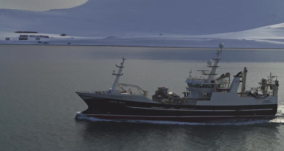 A fishing trawler sails across a calm sea with a snow-covered arctic landscape in the background