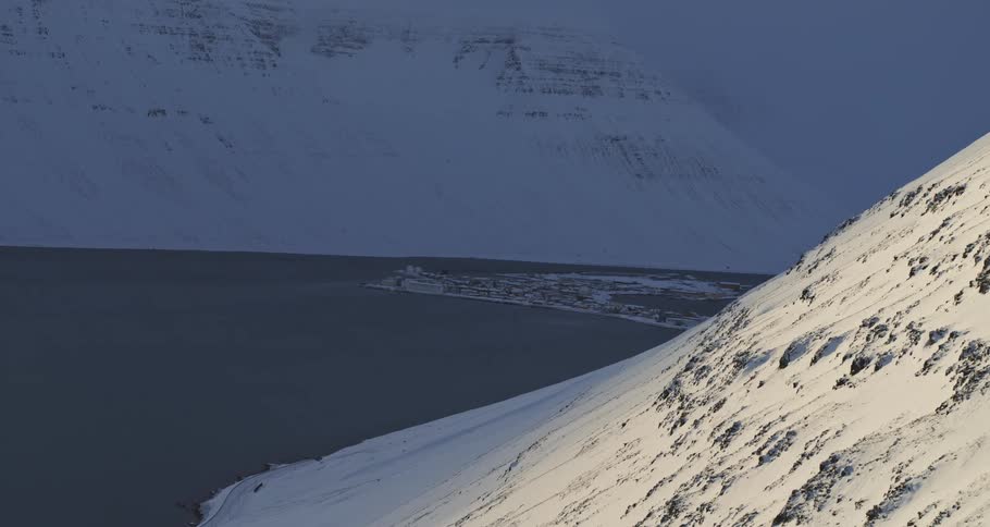 A distant coastal town sits nestled within a massive, snow-covered fjord in the dim light of winter