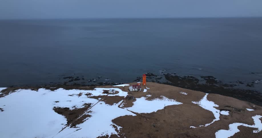 A static aerial shot of a vibrant orange lighthouse on a snowy peninsula overlooking a dark, calm sea on an overcast day