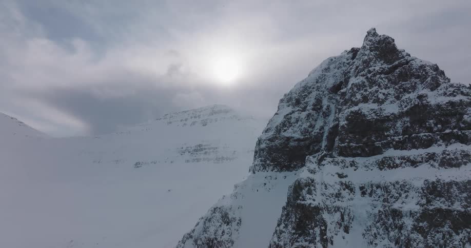 Dramatic aerial reveal of a majestic, snow-covered mountain peak under a hazy sun and cloudy sky