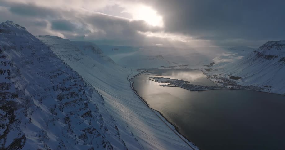 A stunning aerial view captures sunbeams piercing through clouds over a remote Icelandic village nestled between snow-covered mountains and a fjord