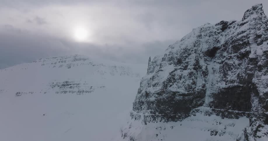 An aerial view pulls back from a dramatic snow-covered mountain peak as a large bird soars through the vast, cold landscape