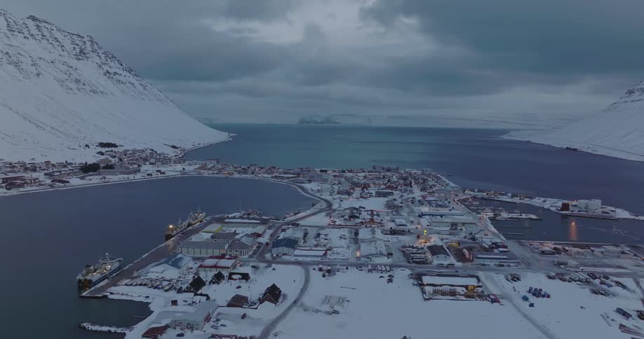 Twilight descends over a remote, snow-covered fishing town and harbor nestled between towering mountains in a wintery fjord