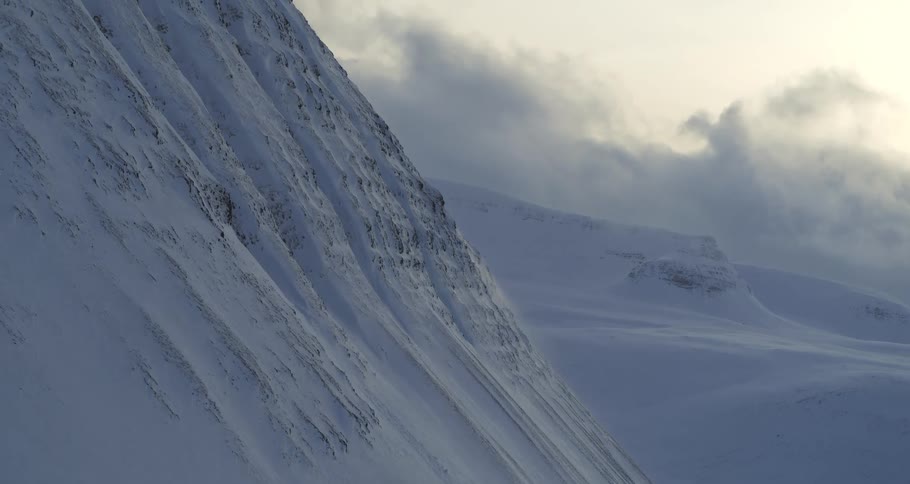 Vast snow-covered mountains stand against a cloudy sky as gusts of wind blow snow across the desolate landscape