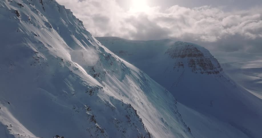 An epic aerial shot follows a rugged, snowy mountainside as powerful winds blow snow across the peaks under a cloudy, sunlit sky