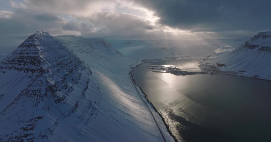 Sun rays break through dramatic clouds to illuminate a majestic, snow-covered fjord in a winter landscape