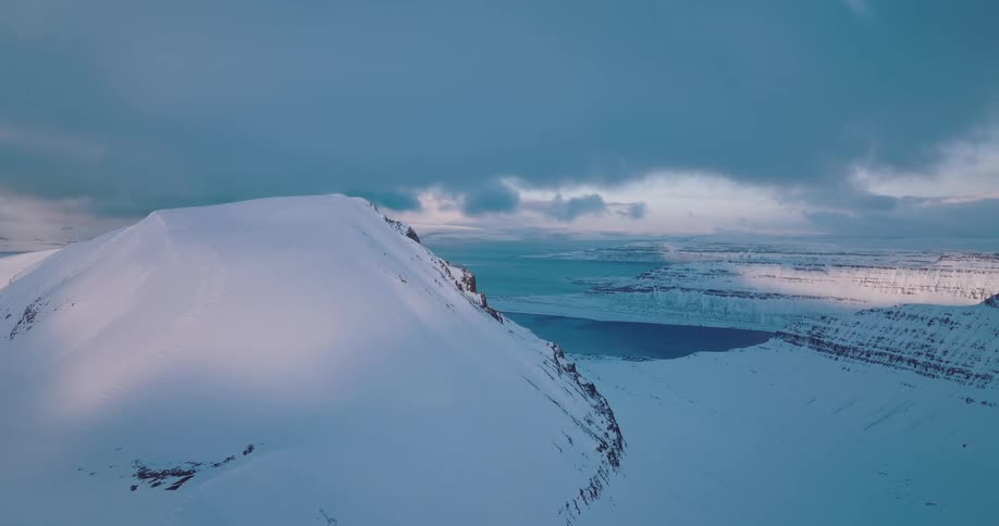 Static aerial view of a majestic, snow-covered mountain overlooking a vast fjord under a dramatic, cloudy sky in winter