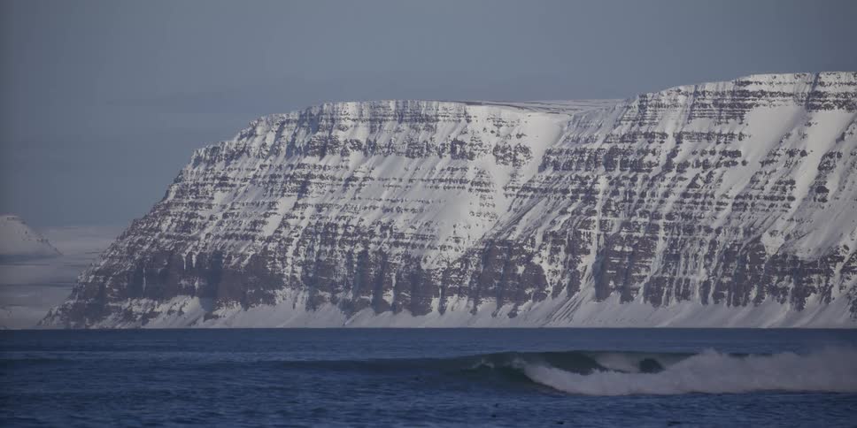 A perfect ocean wave breaks in slow motion in front of a majestic snow-covered mountain