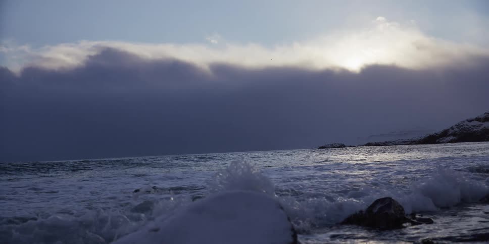 Icy ocean waves crash in slow motion against snow-covered rocks on a cold coastline at dusk