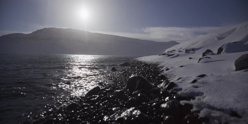 A slow dolly shot along a snowy, rocky shoreline reveals the bright arctic sun reflecting off the calm ocean waves