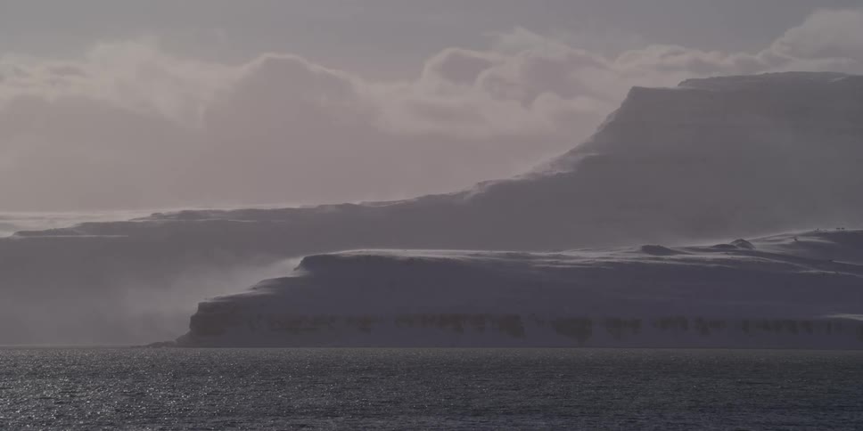 Strong arctic winds whip snow across a desolate, frozen coastline under an overcast sky