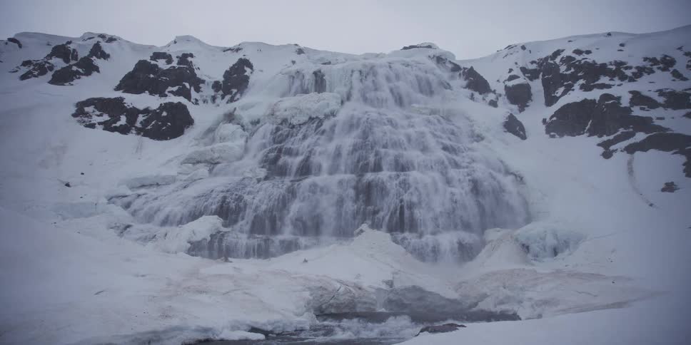 A slow dolly shot reveals the majestic, partially frozen Dynjandi waterfall cascading down a snowy mountain in Iceland during winter