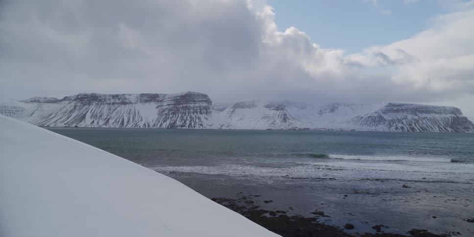 A serene, wide shot of a vast, snow-covered mountain range across a calm ocean fjord on a cloudy winter day