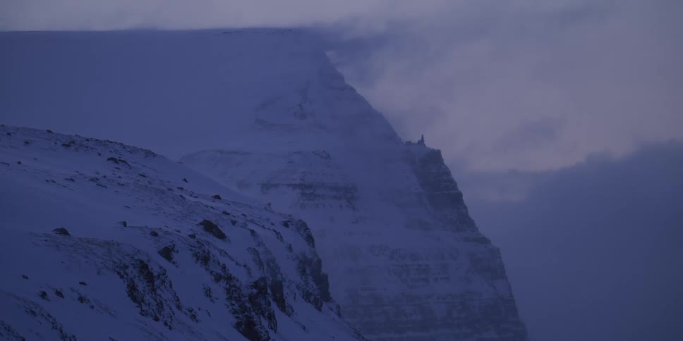 A dramatic, snow-covered mountain peak is engulfed by thick, moving fog in a cold and desolate winter landscape