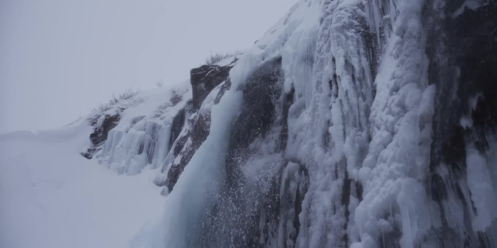 Slow motion captures a majestic waterfall flowing over a snow and ice-covered cliff in a serene winter landscape