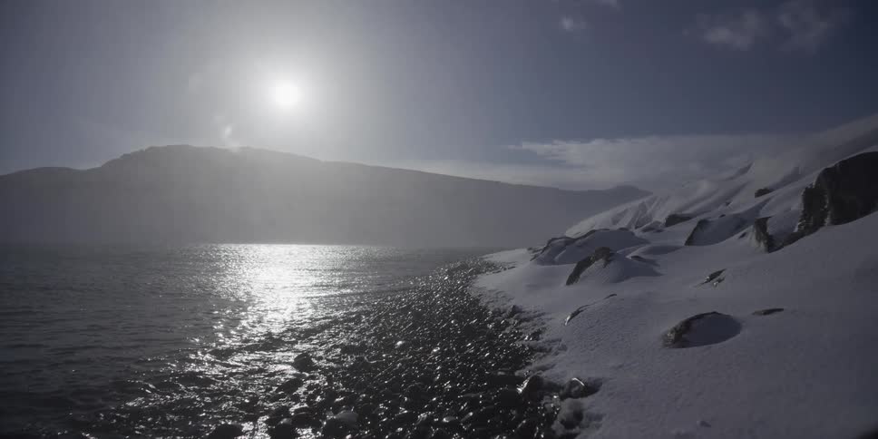 A slow-moving shot reveals the sun reflecting off the water along a cold and rocky snow-covered shoreline