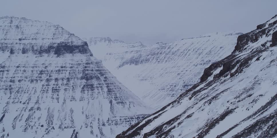 A static wide shot captures the stark beauty of a vast, snow-covered mountain valley under a flat grey sky