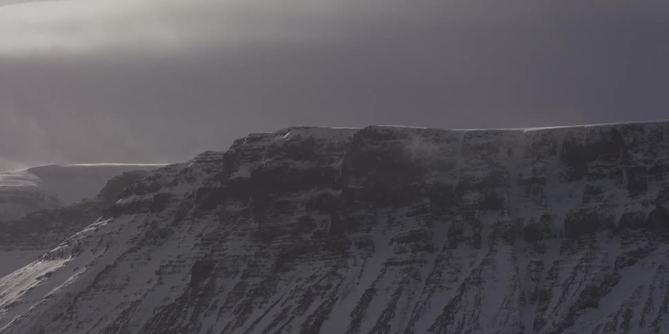Blowing snow whips across the peak of a rugged, icy mountain ridge under a stormy sky