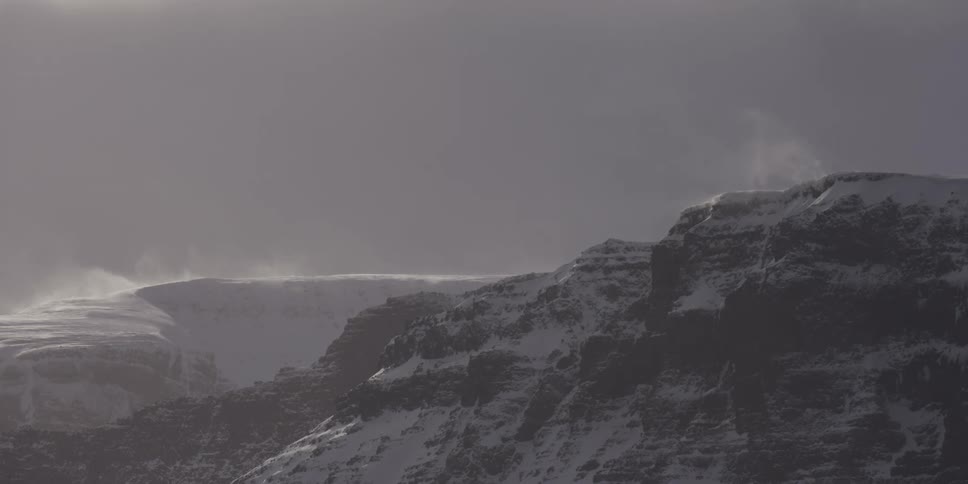 High winds blow snow over the icy peaks of a rugged mountain range in a desolate, frozen landscape