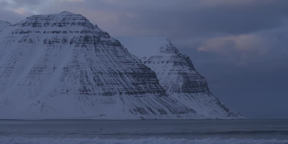 Towering snow-covered mountains meet the cold ocean as waves roll in under a moody, overcast sky