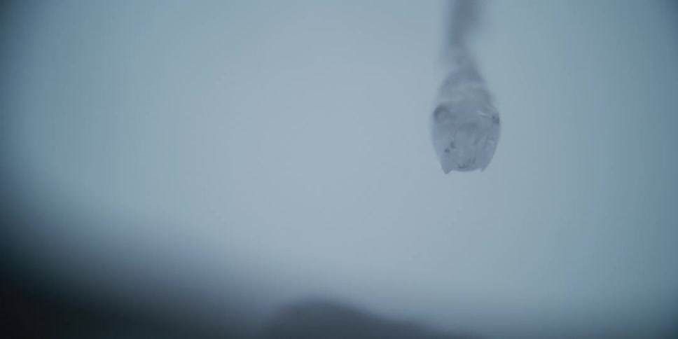 A mesmerizing slow-motion macro view of a single drop of clear water slowly forming and swelling against a soft background