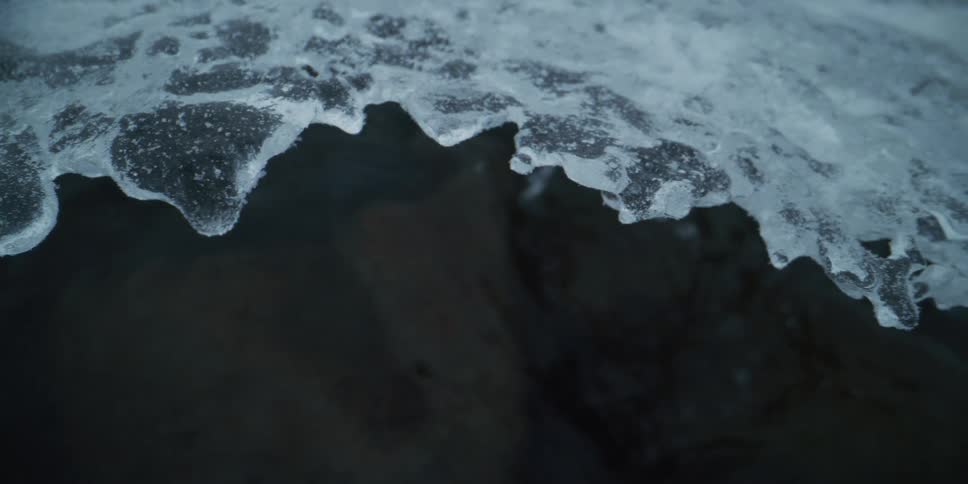 A close-up slow pan reveals the intricate edge of a sheet of ice over a dark, flowing winter stream