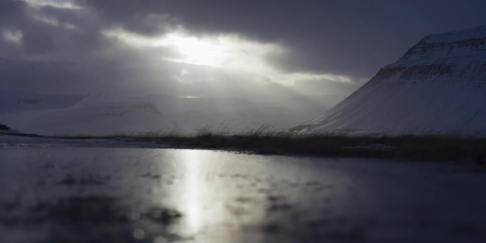 Sunlight reflects off a frozen puddle, illuminating a vast, snowy valley surrounded by dramatic mountains and clouds