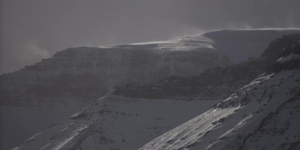 High winds blow snow across a rugged mountain peak during a cold winter storm