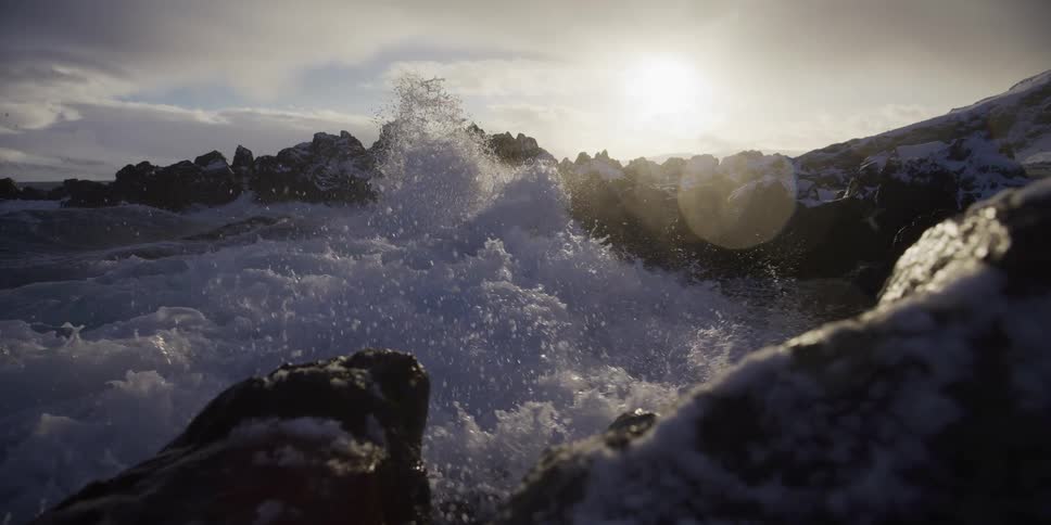 Dramatic slow motion shot of ocean waves crashing over snow-dusted rocks as the sun shines brightly