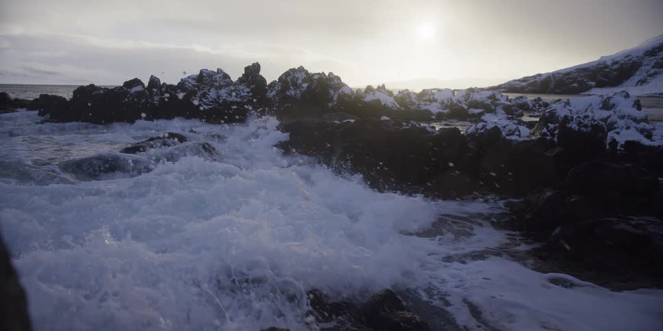 A powerful ocean wave crashes in slow motion against a shoreline of snow-dusted rocks under the winter sun