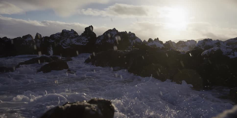 Ocean waves crash over snow-covered rocks in dramatic slow motion as the sun sits low in the sky