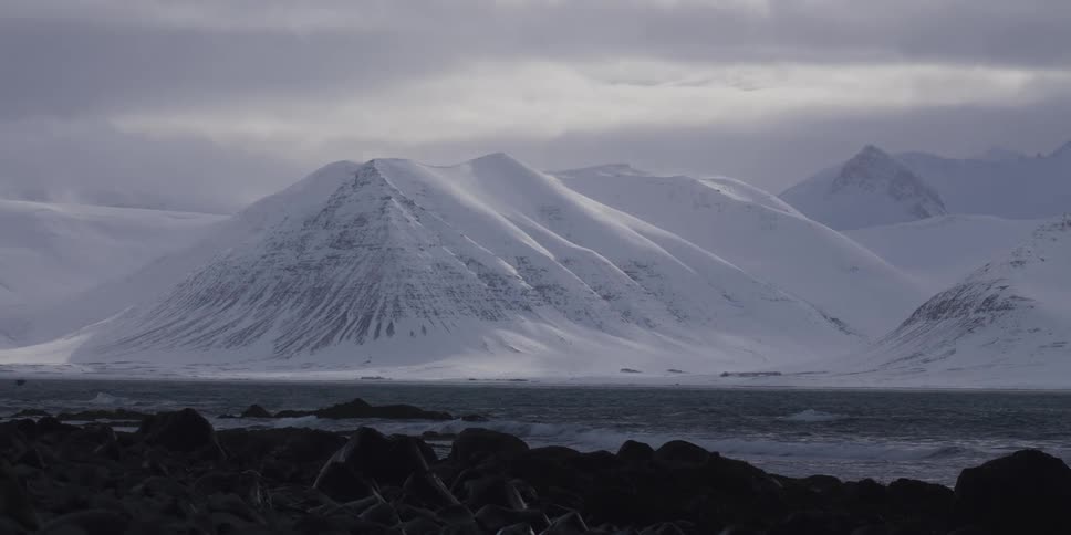 Vast snow-covered mountains rise above the choppy Arctic waters as seen from a dark, rocky shoreline under an overcast sky