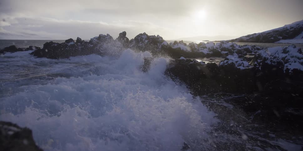 Slow motion shot of a powerful ocean wave crashing against a snowy, rocky shoreline under a pale sun
