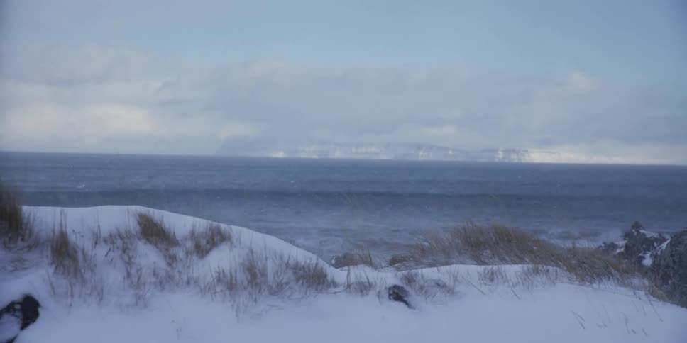 A tracking shot reveals a desolate, snow-covered coastline as strong winds blow snow over the stormy, dark ocean