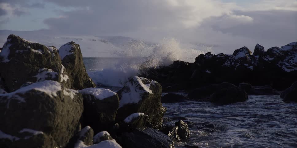 A dramatic slow-motion shot reveals waves crashing on a rugged, snowy coastline with mountains in the background