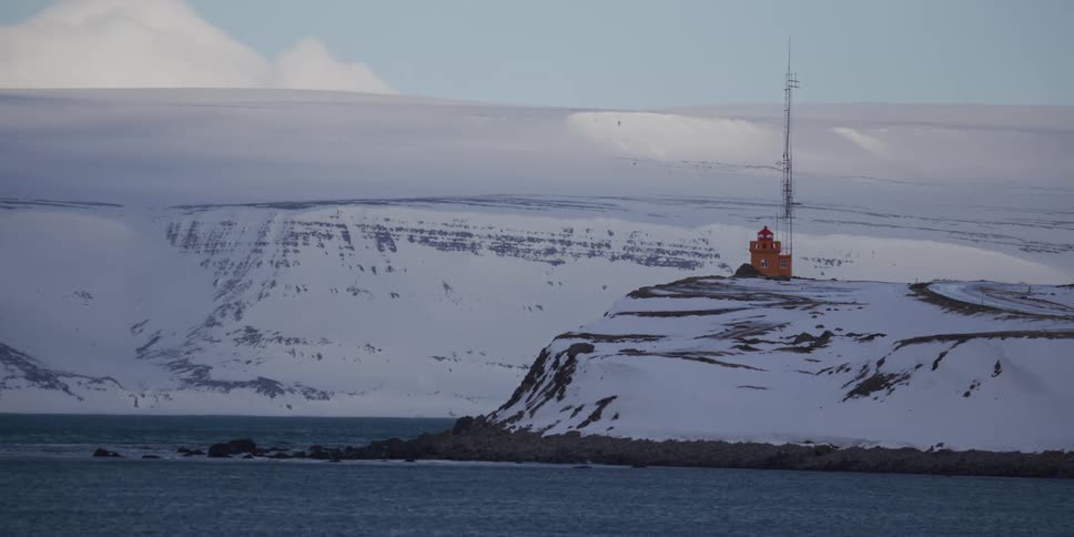 A solitary orange lighthouse and communications tower stand on a snowy cliffside overlooking the water in Iceland