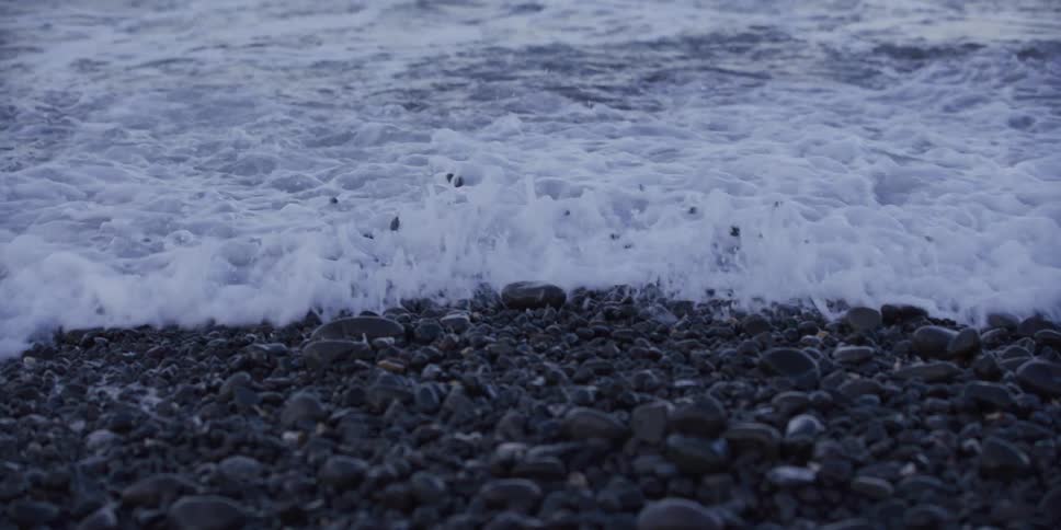 A foamy white ocean wave slowly washes over the dark pebbles of a rocky shoreline