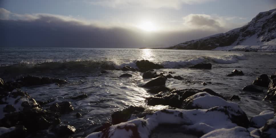 A slow-motion dolly shot captures gentle ocean waves rolling onto a snow-covered rocky coastline at sunset