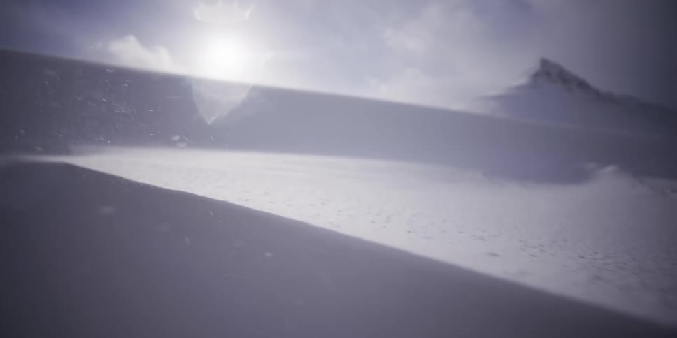 A close-up slow-motion shot captures the wind blowing loose snow across a desolate, sun-drenched mountain landscape