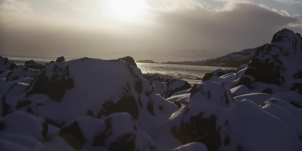 A bright sun shines on the water over a snowy, rocky shoreline on a peaceful winter day