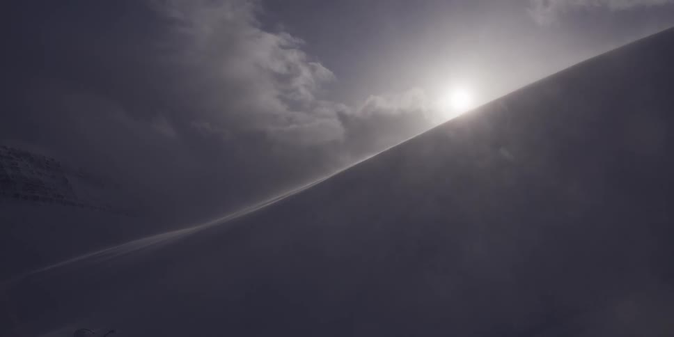 Sunlight pierces through dark clouds as strong winds blow snow across a stark mountain ridge