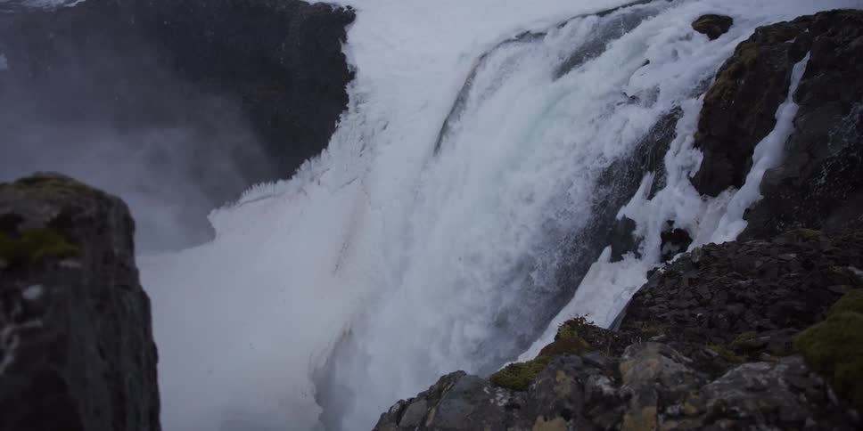 A dramatic slow-motion close-up captures a powerful waterfall cascading over a snow-covered rocky cliff